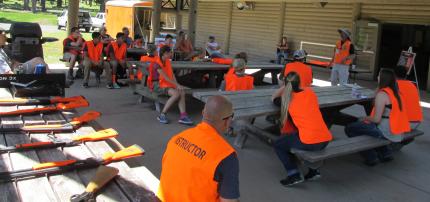 Two hunter education instructors with approximately 20 students in an outdoor setting. All wearing hunter orange vests. Shotguns are lying on a table near one of the instructors.