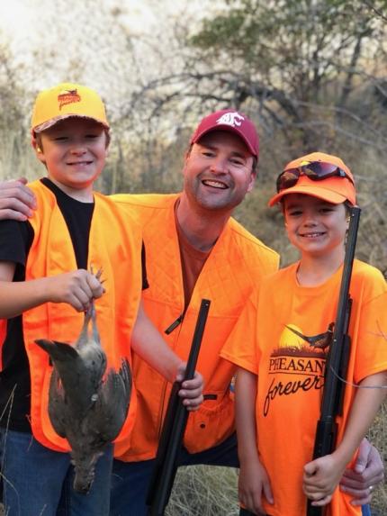 A young boy holding his first harvested partridge smiling with his father and brother. All are dressed in hunter orange.
