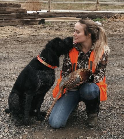 Photo of a hunter holding a harvested pheasant kneeling nose-to-nose with her dog.